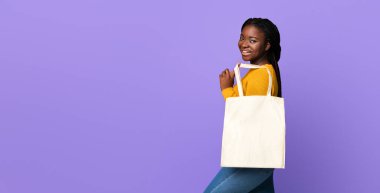 Boş eko çantalı neşeli Afro-Amerikan kadın pozu. Tasarım ya da logonuz için model çanta, mor stüdyo arka planında duran pozitif siyah kadın, kameraya gülümsüyor.