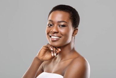 Afrikalı güzellik. Güzel siyah kadın portresi banyo üstüne havlu sarılmış, çenesine dokunuyor ve gri arka planda fotokopi çekerek kameraya gülümsüyor.