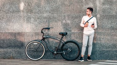 Aktif yaşam tarzı, modern hipster tarzı ve online çalışma. Elinde telefon ve kahve bardağıyla caddede bisikleti olan gri duvarın yanında duran çekici genç adam yan yana bakıyor, panorama, boş alan.