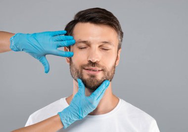 Orta yaşlı, gözleri kapalı bir adam güzellik iğnesi yaptırıyor estetik cerrah ya da hemşire ameliyattan önce hasta yüzüne dokunuyor gri stüdyo arka planı, fotokopi odası. Erkek estetiği kozmetolojisi