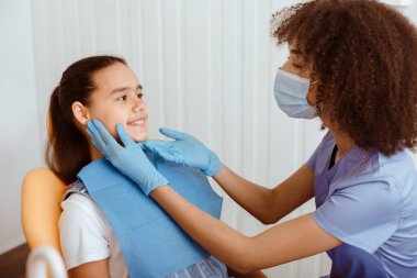Nazik dişçi ve modern çocuk kliniği. Üniformalı genç Afro-Amerikan dişçi koruyucu maske ve eldivenler küçük hastaların ağız bakımı ve diş ağrısını önlemek için kontrol ediyor.