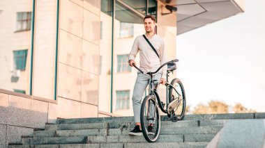 Kendine güvenen genç işadamı şehirde çevre taşımacılığıyla yürüyor, işe gidiyor ve aktif bir hayat sürüyor. Gülümseyen yakışıklı menajer sabah merdivenlerde bisiklet taşıyor, açık havada, boş alanda.