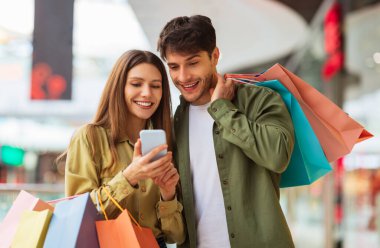Mutlu Çift Alışverişi Cep Telefonu Uygulaması Renkli Alışveriş Çantaları ve Modern Alışveriş Merkezi 'nden Hafta Sonu Kıyafet Alma. Müşteriler Satış için interneti tarayan akıllı telefonları tutuyor. Ticaret.
