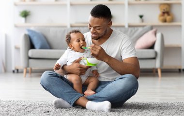 Siyah Baba 'nın sevimli bebeğini kaşıkla beslerken oturma odasında birlikte oturan Afrikalı Amerikalı genç baba, küçük çocukla ilgilenmenin keyfini çıkarıyor.