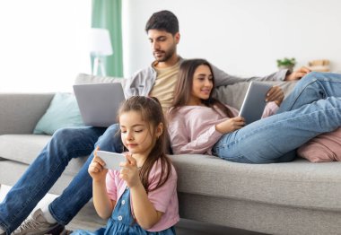 Gadgets bağımlılığı. Doğu ailesi farklı elektronik cihazlar kullanıyor, oturma odasında birlikte dinleniyorlar, ebeveynler kızları yerdeyken kanepede oturuyorlar. Modern teknolojiler, phubbing kavramı