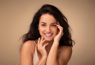 Bej renkli stüdyo arka planında poz veren, güzel, esmer bir kadının yakın plan portresi. Yüzüne dokunuyor ve gülümsüyor. Güzellik tedavisinden sonra pürüzsüz cildinin tadını çıkarıyor.