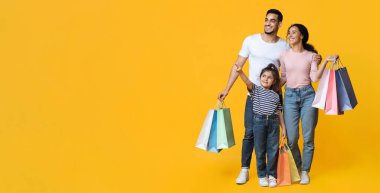 Alışveriş Teklifi. Küçük kızıyla gülümseyen bir Arap ailesi alışveriş torbalarını taşıyorlar ve sarı arka plan, mutlu ebeveynler ve sezonluk satışlardan hoşlanan kadın çocuk Panorama 'yı işaret ediyorlar.