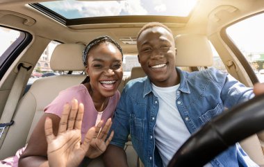 Mutlu Afro-Amerikan çifti araba gezisi yaparken selfie çekiyor, arabada oturuyor, kameraya sarılıyor ve neşeyle gülümsüyor, binlerce yıllık siyahi kadın erkek arkadaşına sarılıyor ve el sallıyor, güneş parlıyor.