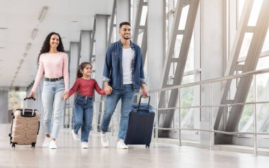 Havalimanı koridorunda bavullarıyla yürüyen mutlu bir Orta Doğulu aile, neşeli bir Arap ebeveyni ve küçük kızı birlikte yurtdışına seyahat ediyor, el ele tutuşuyor ve gülümsüyor, boşluğu kopyalıyor.