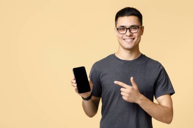 Blogcu müşterilere daha iyi bir teklif sunmakta ısrar ediyor. Kahverengi tişörtlü ve gözlüklü mutlu genç adam, boş ekran reklam uygulamalı cep telefonunu işaret ediyor, bej arka planda izole edilmiş.