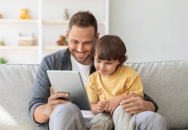 Çocuk gelişimi içeriği. Genç, pozitif baba, sevimli oğluna dijital tablet gösteriyor. Çocuk internette eğitim programından zevk alıyor. Evdeki kanepede oturuyor.