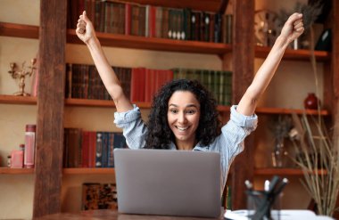 Evet, ben bir kazananım. Heyecanlı duygusal mest olmuş bir bayan, başarıyı, zaferi ya da harika haberleri kutluyor, masada oturuyor, kişisel bilgisayarını kullanıyor, evet diye bağırıyor ve ellerini kaldırıyor. Gülümseyen mutlu kadın neşe içinde