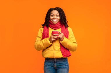 Kışlık ceketli, ceketli, elinde kahve fincanı olan siyah bir kadın sarı stüdyonun arkasında kameraya gülümsüyor. Afro-Amerikan Kadın Sıcak İçecekten Hoşlanıyor. Sonbahar İlan Pankartı