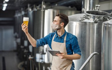Bira kalite kontrol. Önlüklü ve koruyucu maskeli dijital tabletli bir adam bira fabrikasında bir bardak biraya bakıyor.