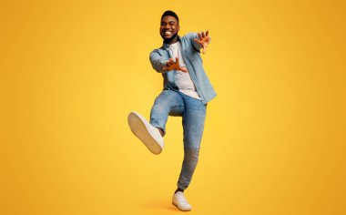 Komik Afrikalı Amerikalı adam dans ediyor ve ellerini sarı stüdyo arka planında kameraya uzatıyor.