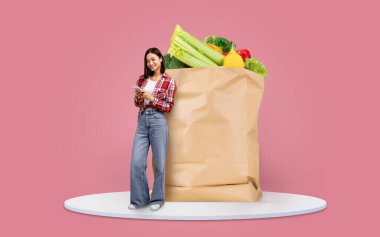 Bir müşteri, akıllı telefonundaki listeyi kontrol ederken içi taze sebze ve meyve dolu büyük bir kesekağıdının yanında duruyor. Market alışverişi önemli bir günlük faaliyet haline geldi..
