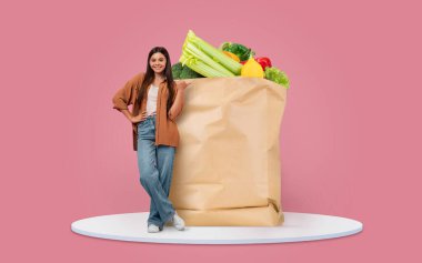 Neşeli bir birey taze sebze ve meyvelerle dolu büyük bir kesekağıdının yanında duruyor. Bu durum, market alışverişi ve modern yaşam tarzlarında ev teslimatı eğilimini yansıtıyor..