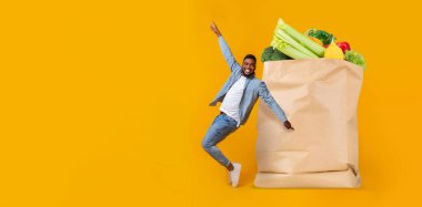 Sıradan giyinmiş mutlu bir müşteri, marketten alışveriş yaptıktan sonra kutlama yapar. Parlak sarı arka plan, kağıt torbadaki sebzelerin tazeliğini arttırır..