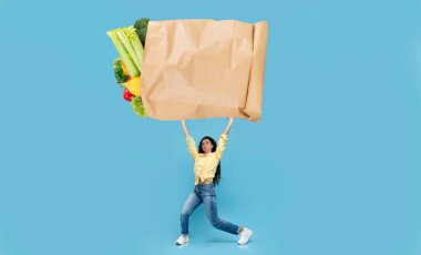 Bir müşteri gururla canlı sebzeler ve meyvelerle dolu büyük bir kesekağıdı tutar. Arkaplanda, market alışverişinin heyecanını vurgulayan açık mavi bir renk yer alıyor..