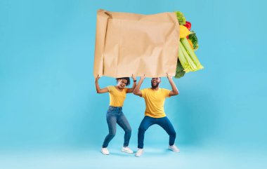 Birbirine uyumlu sarı gömlekler giyen bir çift, taze sebzelerle dolu büyük bir kesekağıdını kaldırırken gülümsüyor. İzolasyon ortasındaki yeni market alışverişini temsil ediyorlar..