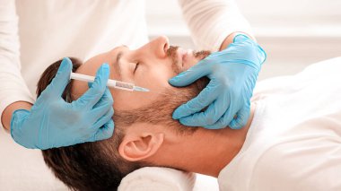 Kozmetik uzmanı klinikte orta yaşlı yakışıklı bir adam için yaşlanma karşıtı işlem yapan mavi lastik eldivenleri verdi. Sakallı işadamı kuaförde güzellik iğnesi yaptırıyor.