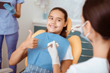 Modern klinikte tıbbi ekiple hızlı ve acısız diş tedavisi. Sandalyedeki gülümseyen İspanyol çocuk, bin yıllık beyaz önlüklü, koruyucu maskeli ve lastik eldivenli doktora baş parmağını gösteriyor.