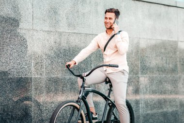 Acil çağrı, hareket ve aktif çalışma, spor yaşam tarzı. Gülümseyen çekici milenyum adamı menajeri telefonda konuşuyor ve şehirde bisiklet sürüyor, gri sokak duvarında arka planda, açık havada, serbest alanda.
