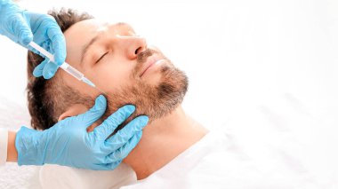 Sakallı, orta yaşlı bir adam güzellik salonunda güzellik iğnesi yaptırıyor. Kozmetik uzmanı klinikteki yakışıklı adam için yaşlanma önleyici prosedür uygulaması yapan mavi lastik eldivenleri teslim etti. Yüz bakımı, erkek kozmetoloji konsepti.