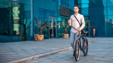 Modern işçi ve kahve molası. Çantalı ve kahveli adam, bisikletle ofis binasının yakınına gidiyor.