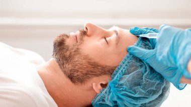 Saç filesi takmış sakallı iş adamının estetik kliniğinde yaşlanma önleyici doldurma ameliyatı, yan görüş. Koruyucu mavi eldivenli plastik cerrah orta yaşlı adamın alnına hyaluronik asit enjekte ediyor.