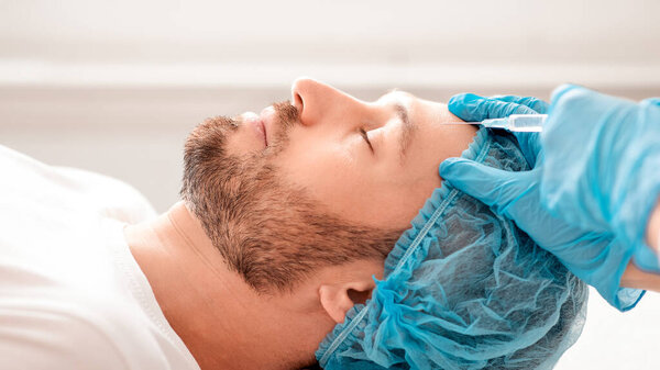 Крупный план бородатого бизнесмена в сетке для волос, получающего наполнитель против старения в эстетической клинике, вид сбоку. Пластический хирург в защитных голубых перчатках вводит гиалуроновую кислоту в лоб мужчины средних лет
