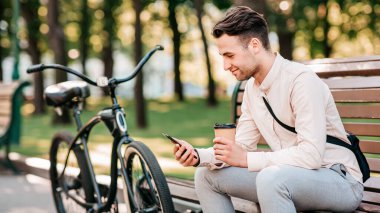 Dışarıda çalış, biraz ara ver ve sabah ofise git. Gülümseyen milenyum adamı menajer bisikletle bankta oturur, bir fincan kahve alır ve açık havada, şehirde, araçta mesaj okur.