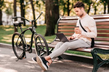 Zevkle çalışın. Yandan bakan yakışıklı adam, günlük kıyafetlerini giyiyor, dizüstü bilgisayarda yazıyor ve kahve ısmarlıyor, şehir parkında, açık havada, bisikletin yanında oturuyor.