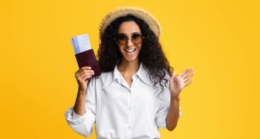 Yaz şapkalı, gözlüklü, pasaportu ve biletleri olan neşeli genç bayan, seyahate çıkmaya hazır olan heyecanlı Brunetter hanımı, sarı stüdyonun arka planında duruyor ve kameraya bakıyor,