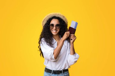 Pasaportu ve biletleri sarı arka planda olan, yaz şapkası ve gözlük takan, tatile çıkmaya hazır neşeli bir bayan, kameraya bakıp gülümseyen,
