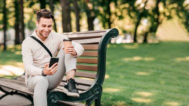 Genç bir adam canlı bir şehir parkındaki bankta dinleniyor, bir fincandan kahve yudumluyor ve akıllı telefonuna bakıyor. Güneş ağaçların arasından parlıyor, canlı bir atmosfer yaratıyor, uzayı kopyalıyor.