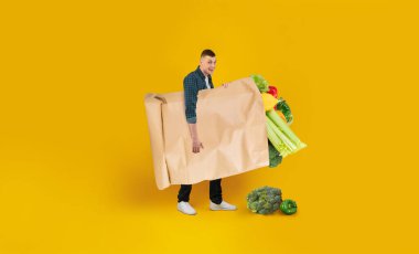 Bir insan, taze sebzeler de dahil olmak üzere çeşitli sebzelerle dolu büyük bir kesekağıdını tutarken gülümsüyor. Canlı sarı arka plan market alışverişinin neşeli atmosferini arttırır..