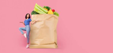 Bir müşteri, içi taze sebze, marul, brokoli ve biber dolu büyük bir kese kağıdının yanında duruyor. Bu, market alışverişinin ve teslimatın yeni normalini vurguluyor.