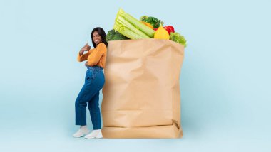 Taze sebze ve meyvelerle dolu büyük bir kesekağıdının yanında duran bir müşteri, yeni normal market alışverişini ve eve teslimatını kucaklıyor..