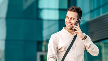 Genç bir adam akıllı telefonuyla konuşuyor, neşeli bir şehir caddesinde bisikletinin yanında dururken gülümsüyor. Modern binalar arka planda şehir yaşamını, kopyalama alanını yansıtıyor.