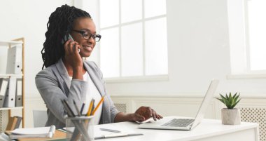 İş İletişimi. Afro kadın modern ofiste dizüstü bilgisayar kullanarak cep telefonu konuşuyor. Kopyalama Alanı ile Panorama