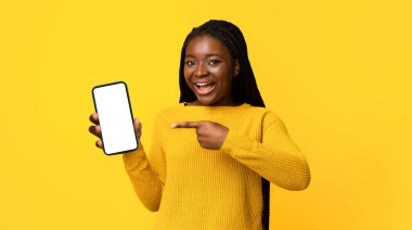 Gülümseyen Afrikalı Amerikalı Kadın Boş Siyah Akıllı Telefon ekranına işaret ediyor, Mokup Tasarımınız, Tavsiye Uygulamanız ya da Web siteniz için bedava yer gösteriyor, Stüdyoda Sarı Arkaplanda Poz Veriyor