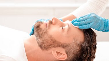 Tanınmayan kozmetik uzmanının, pamuk pedi kullanarak toner ile sakallı adamı temizleyişinin yan görüntüsü. Orta yaşlı iş adamının kaplıcada güzellik prosedürleri var.