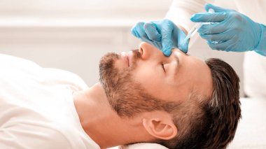 Modern kuaförde sakallı bir adamın güzellik iğnesi yaptırışının yan görüntüsü. Kozmetik uzmanı, yakışıklı bir adam için yüz doldurma işlemi için mavi lastik eldivenleri teslim etti. Yüz bakımı, erkek kozmetoloji konsepti.