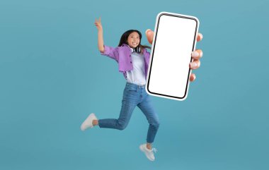 Heyecanlı Asyalı kadın, mavi stüdyo arkaplanı, kolaj, tüm vücut uzunluğu, afiş üzerinden atlarken boş akıllı telefon ekranı gösteriyor. Mutlu kadın havalı mobil uygulama ya da web sitesi öneriyor