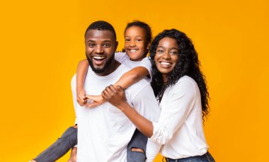 Aile eğlencesi. Genç Afrikalı Amerikalı ebeveynler küçük kızlarıyla vakit geçiriyorlar, sarı arka planda birlikte gülüyorlar.