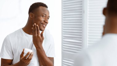 Erkek Cilt Bakımı. Siyah adam nemlendirici losyon kremi kullanıyor cilt bakımını yapıyor evdeki banyoda aynanın yanında duruyor. Erkek Güzelliği ve Kişisel Bakım Rutini. Panorama