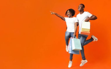 Duygusal Afro-Amerikalı çift, alışverişleri yükseliyor ve neşeli bir şekilde boş alanı işaret ediyor, turuncu stüdyo arka planı. Reklam peşinde koşan mutlu siyah adam ve kadın
