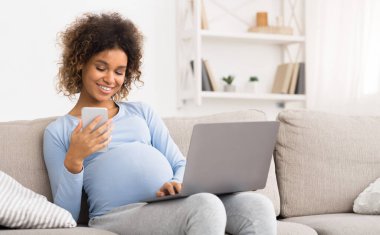 Diz üstü bilgisayar ve cep telefonu kullanan, koltukta dinlenen hamile Afrikalı-Amerikalı kız.