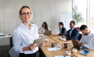 Başarılı iş kadını HR Dijital Tablet Tutarken Modern Ofiste Çeşitli İş Adamları ile Masa Başında Duran Kameraya Gülümsüyor. İş ve İstihdam, İnsan Kaynakları. Boş Alan